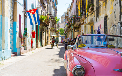 Imagen de una calle típica de Cuba con su bandera, gente trancitandola y un auto de época, típico del pais, de color rosa.